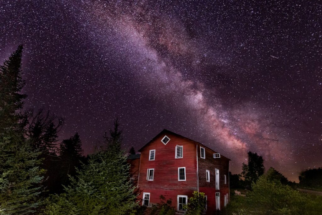 An old red wooden building surrounded by trees, beneath the night sky full of stars 