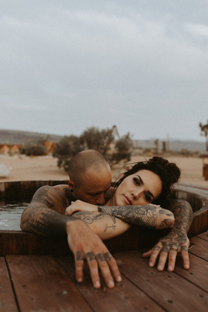 A couple, both with light skin and lots of tattoos, sitting in a hot tub in the desert. The man is kissing the woman's shoulder.