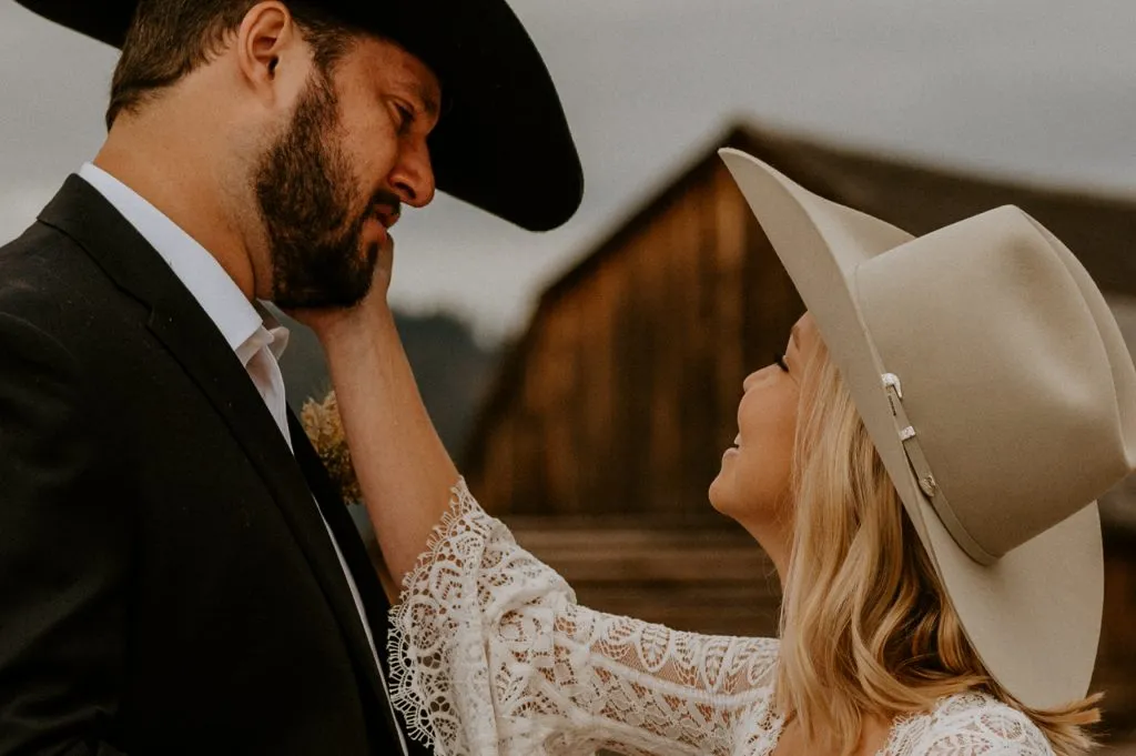 A man with a dark beard, wearing a suit and a black cowboy hat, and his wife, who has long blonde hair and is wearing a lace wedding dress and beige cowboy hat. The wife has her hand on her husband's cheek, and he appears to be crying.