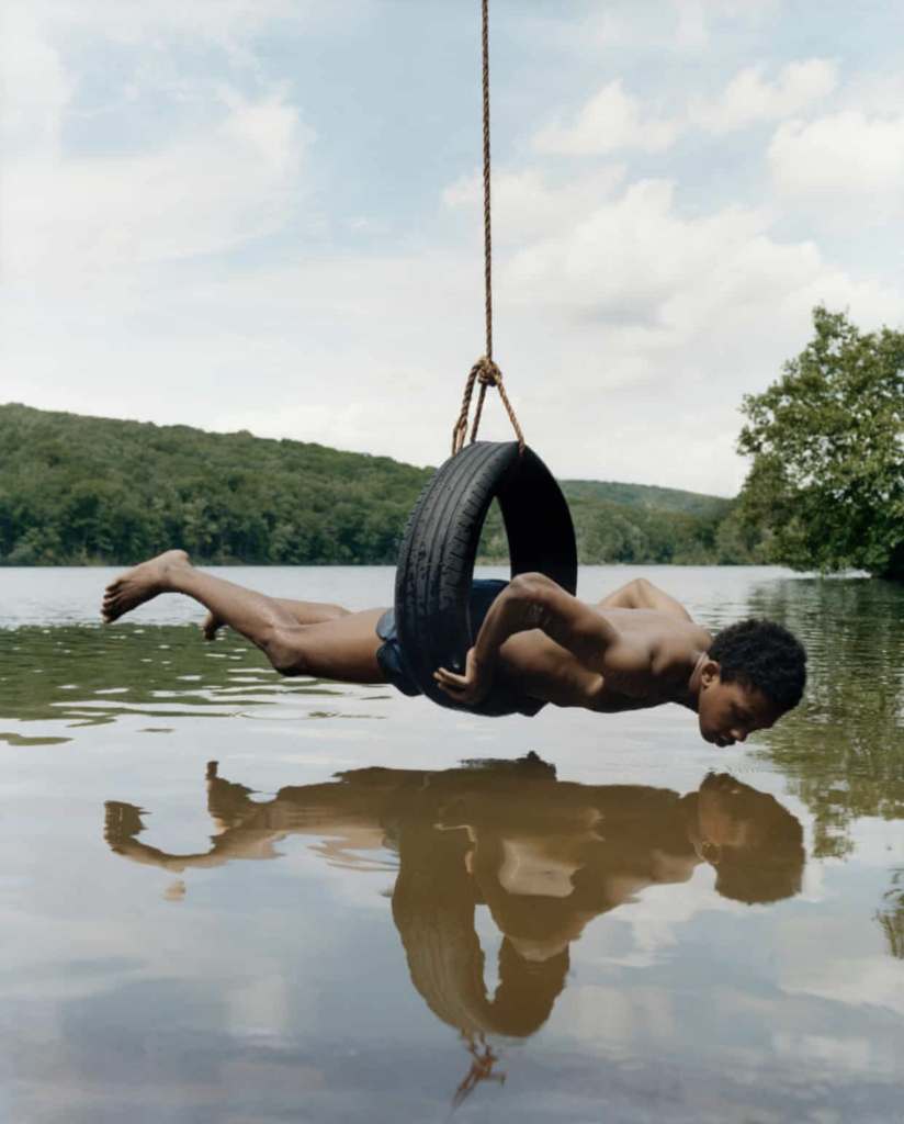A young black man in a tire swing suspended over a lake, looking into his own reflection.