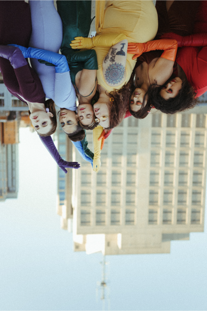 an upside-down photograph of six women, each dressed in a different colour of the rainbow, leaning on each other and extending their arms toward the sky. There is a tall office building in the background.