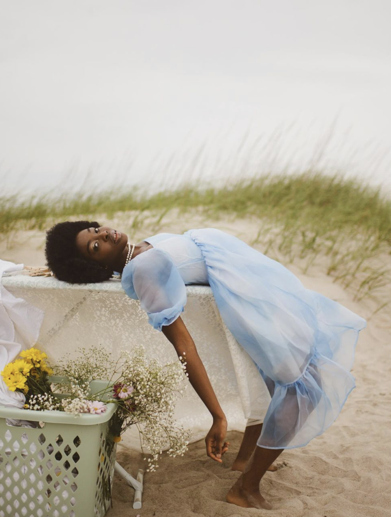 A woman with dark skin and dark hair, wearing a light blue dress, bent backwards over an ironing board that is set up on a beach. There is a laundry bin with flowers in it placed next to her.