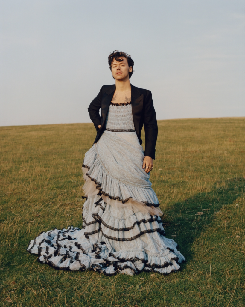 Portrait of Harry Styles. A man with light skin, brown hair, and chest tattoos, wearing a light blue floor-length gown and a dark blazer, standing in an empty field.