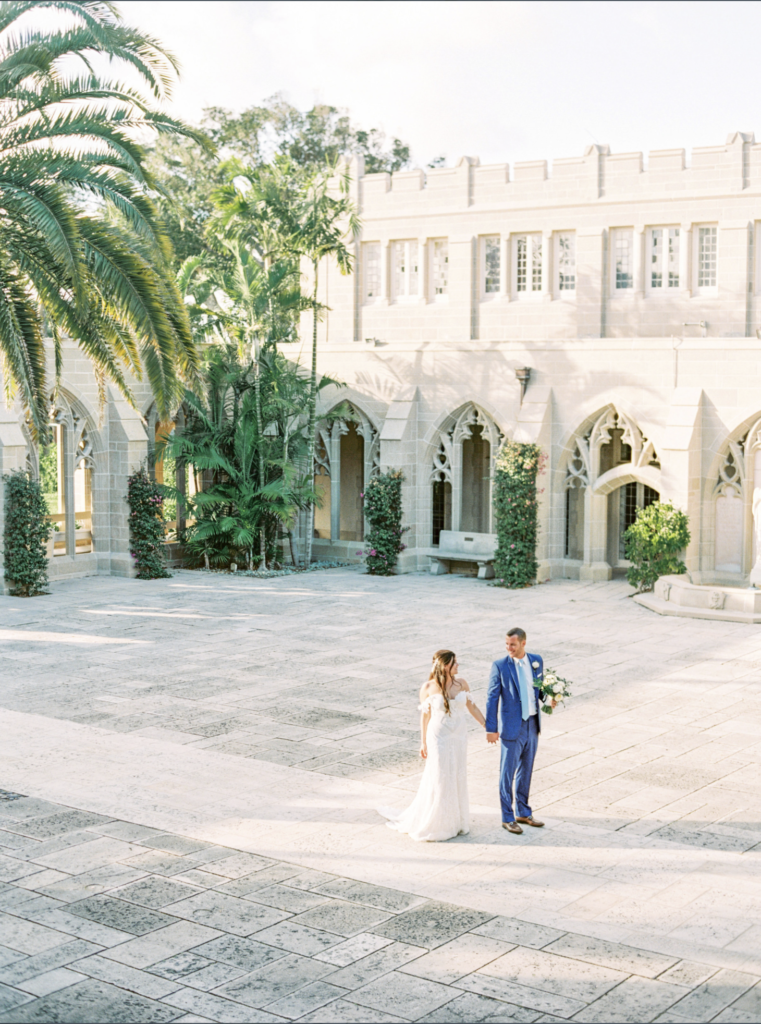 A wide shot of a couple in wedding attire holding hands in the courtyard of an old stone church, with palm trees around the perimeter.