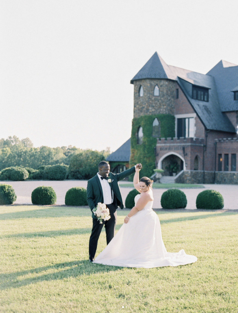 A couple in wedding attire outside of a large red brick estate. The man is twirling his bride on the lawn.