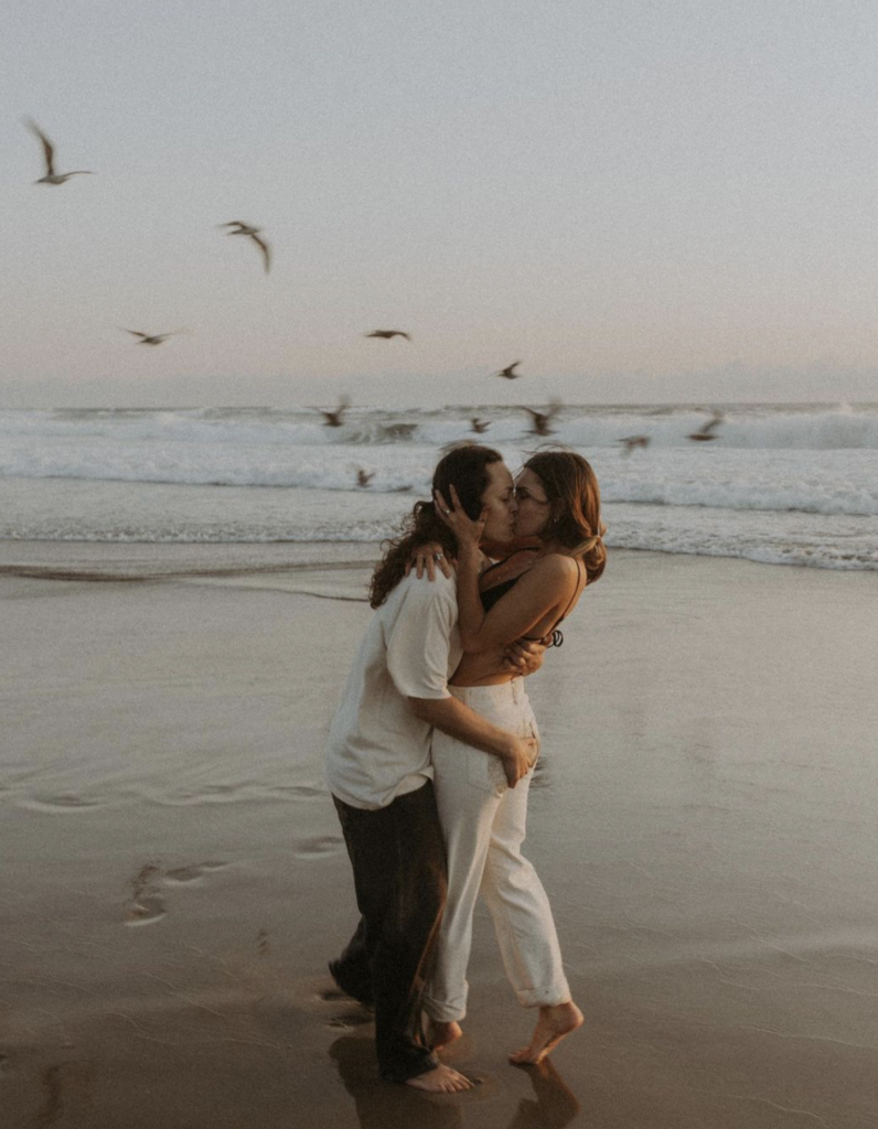 Two women, with tan skin and dark hair, hugging and kissing on a beach at sunset. There is a flock of birds flying behind them.