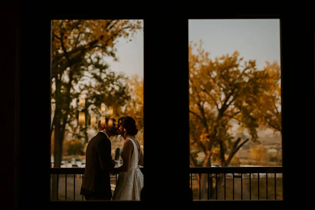 A newlywed couple shares a kiss on a balcony. The photo is shot through a window, framing the scene.