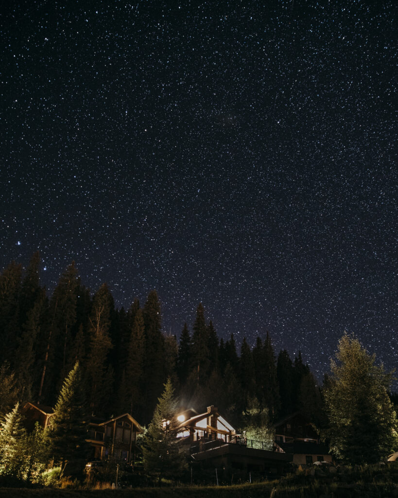 The night sky, full of stars, above a forest and a cabin lit from within in the foreground.